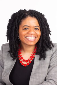 Headshot of woman with black hair, gray blazer, black shirt, and red beaded necklace