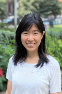 a woman with medium-length black hair wearing a white top, standing outdoors with a blurred garden background.