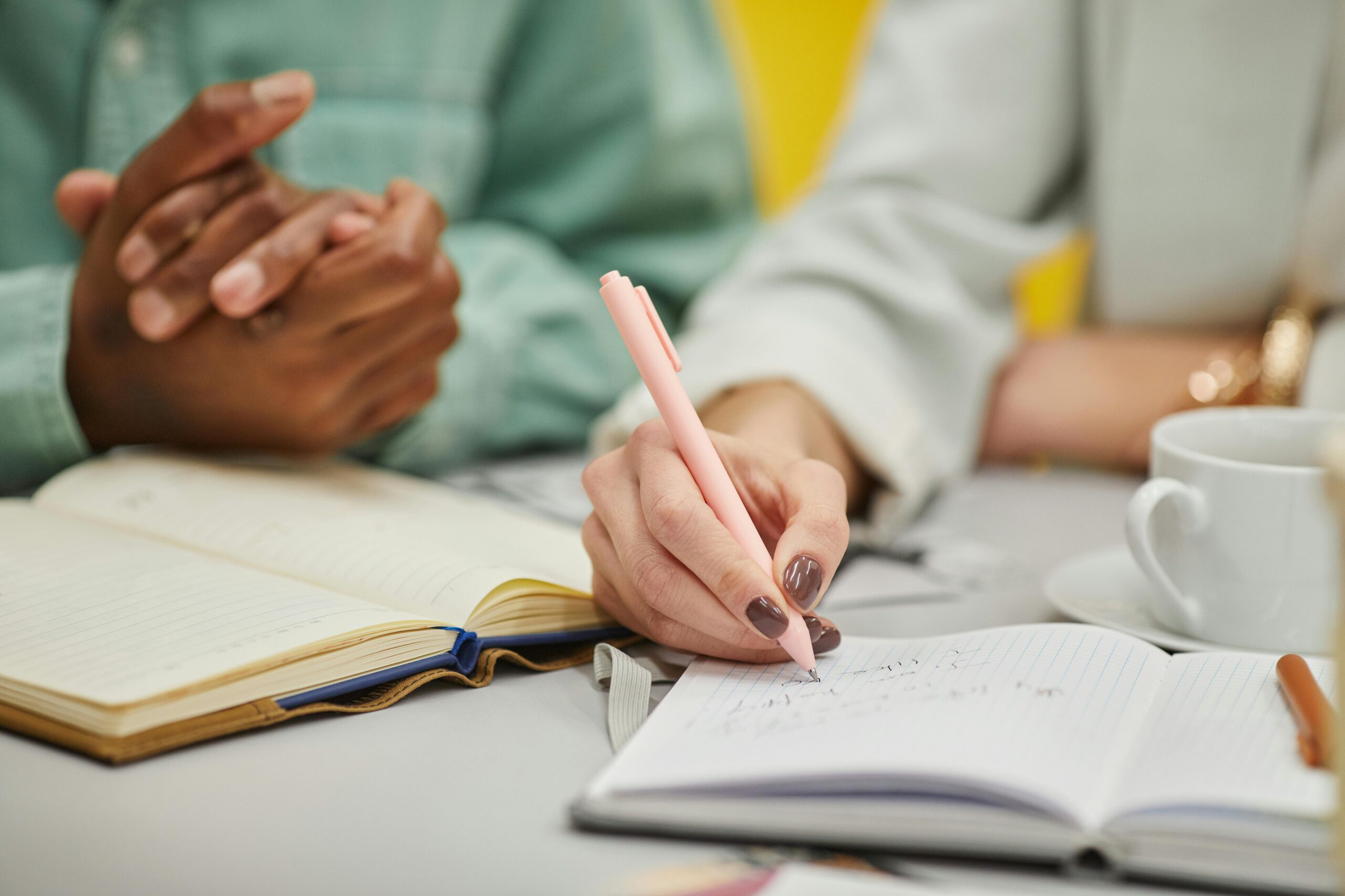 Close-up of two people taking notes and discussing at a table with books and coffee.