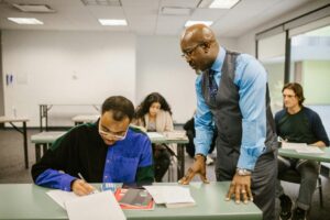 A teacher in a blue shirt and vest leans over to assist a student who is writing in a classroom, with other students working in the background.