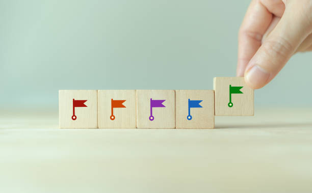 Hand placing a wooden block with a green flag icon next to four other blocks with red, orange, purple, and blue flags, symbolizing milestones or progress.