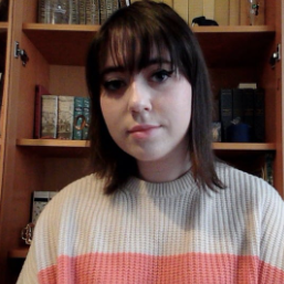 A woman with dark hair and bangs wears a cream and pink sweater while sitting in front of bookshelves.