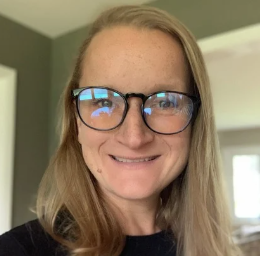 A woman with long blonde hair, glasses, and a black top smiles indoors near a window.