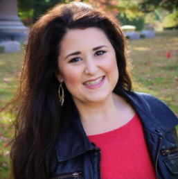 A woman with long dark hair smiles outdoors while wearing a black leather jacket and red top.