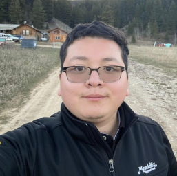 A man wearing glasses and a black jacket stands outside on a dirt path with trees and cabins behind him.