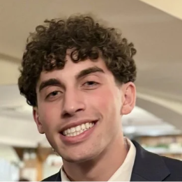 A young man with curly hair and a suit jacket smiles widely indoors.