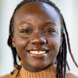 A woman with braided hair and a brown sweater smiles brightly in front of a blurred background.