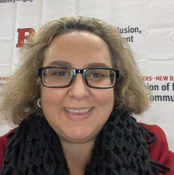 A woman with curly blonde hair, glasses, and a black scarf smiles in front of a Rutgers backdrop.