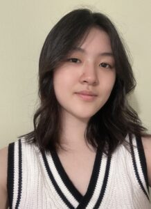 A young woman with shoulder-length dark hair wears a sleeveless white knit top with black trim and stands against a light-colored wall.