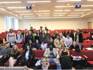 A large group of students and their instructor pose together in a lecture hall, smiling in front of rows of red auditorium seats.