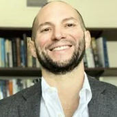 A man with a beard wearing a white shirt and blazer smiles in front of a bookshelf.