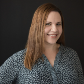 A woman with straight brown hair wearing a patterned blouse smiles in front of a black background.