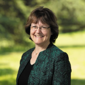 A woman with short brown hair wearing glasses and a green-patterned blazer smiles outdoors in a park setting.