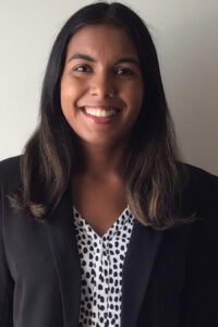 A woman wearing a black blazer and patterned blouse smiles while standing in front of a plain light-colored wall.