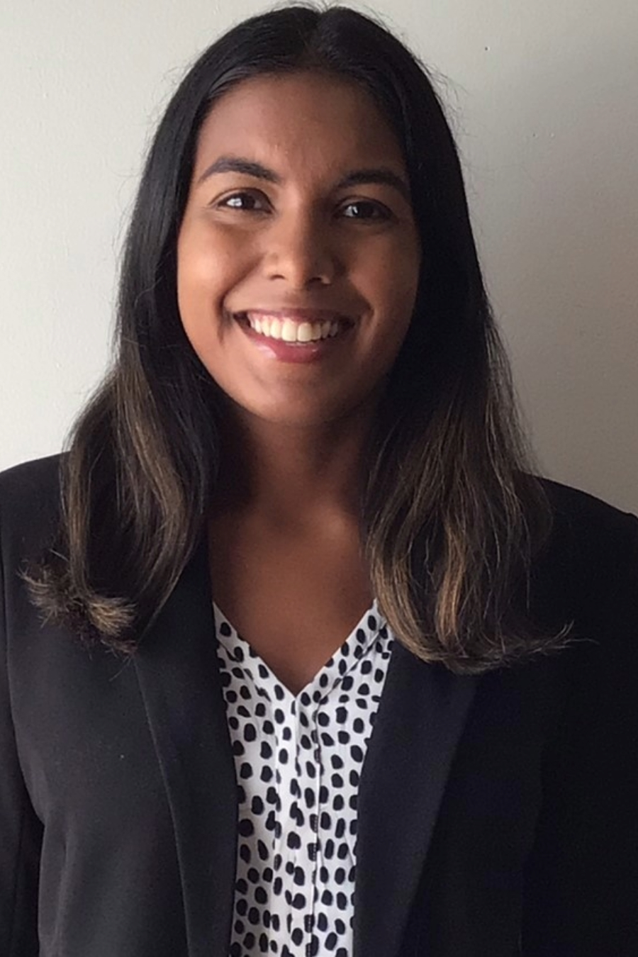 A woman wearing a black blazer and patterned blouse smiles while standing in front of a plain light-colored wall.
