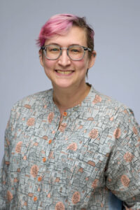 A person with short pink hair, glasses, and a patterned shirt smiles in front of a plain background.