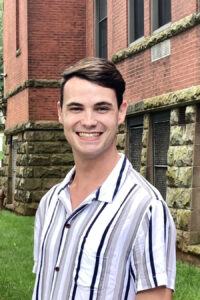 A man wearing a striped short-sleeve shirt smiles while standing outside in front of a brick building.