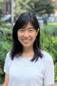 a woman with medium-length black hair wearing a white top, standing outdoors with a blurred garden background.