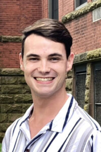A man wearing a striped short-sleeve shirt smiles while standing outside in front of a brick building.