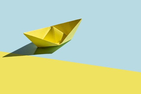 A yellow paper boat sits on a yellow and blue background, casting a shadow.