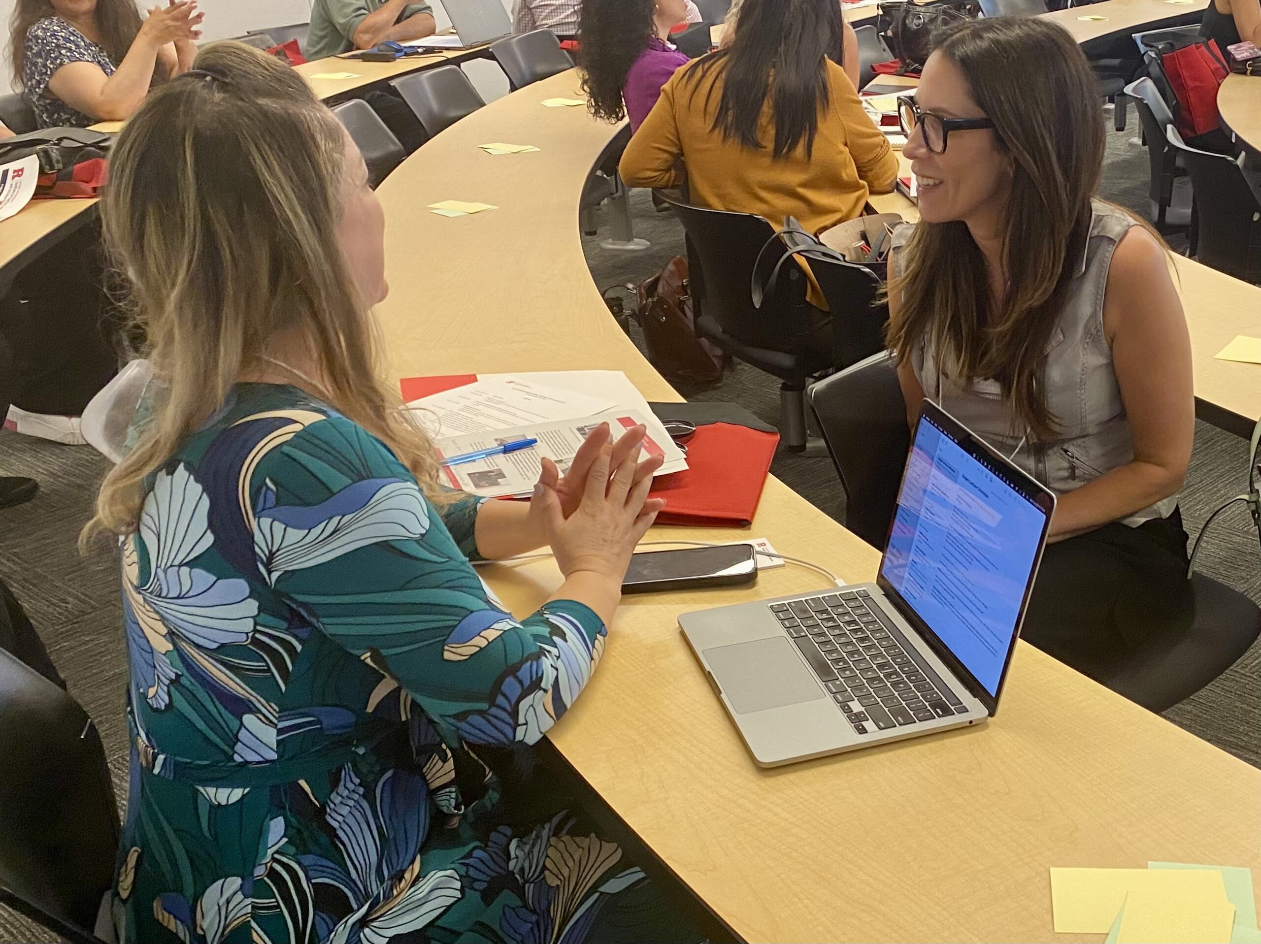 Two women seated across from each other in a classroom engage in conversation during a breakout session, with open laptops and documents on the desk.