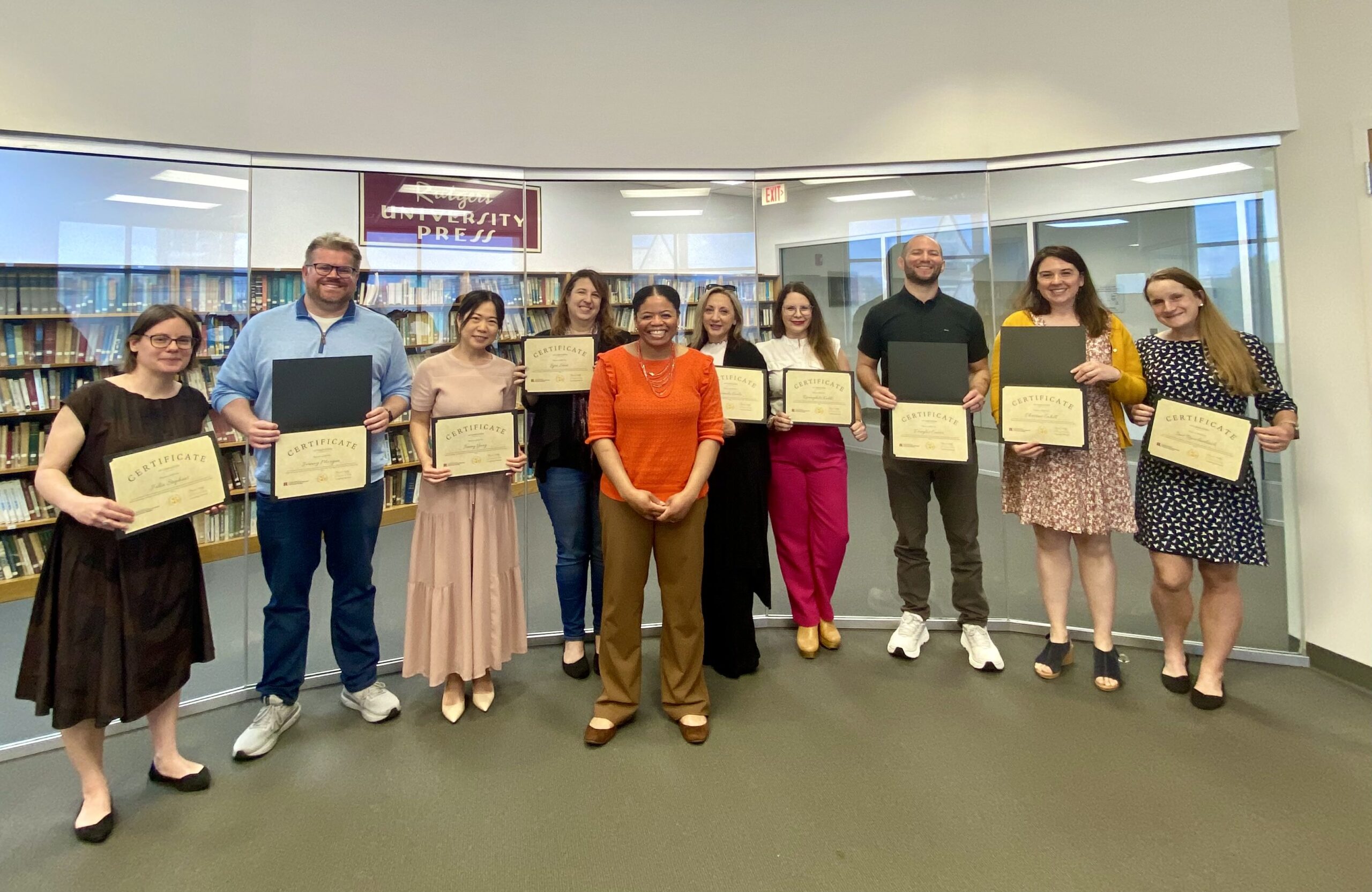 A group of people stand in a library holding certificates, smiling alongside a facilitator in an orange top, with a Rutgers University Press sign visible in the background.