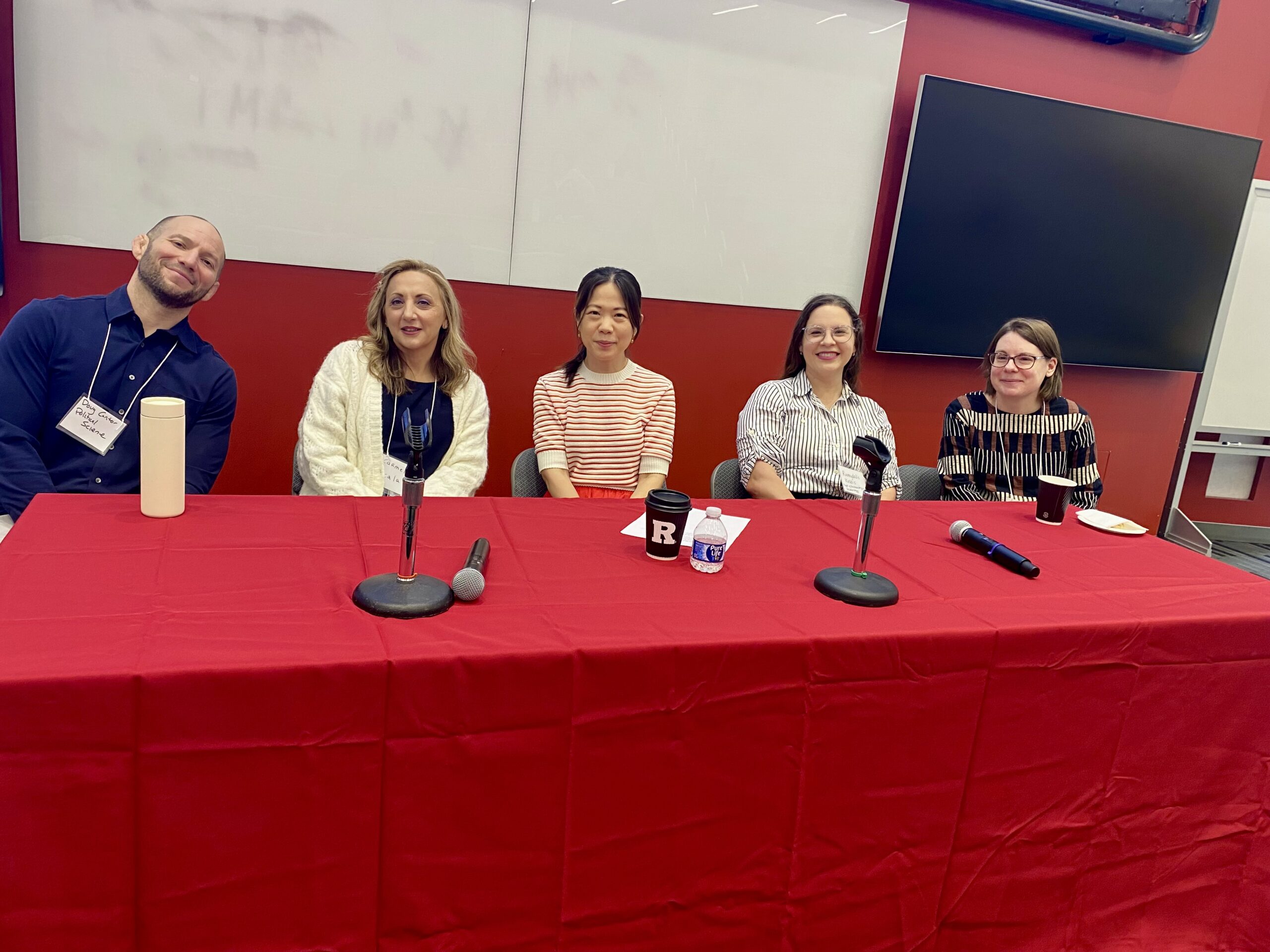 Five panelists sit at a red table with microphones in front of them, smiling and ready for a discussion in a lecture hall.