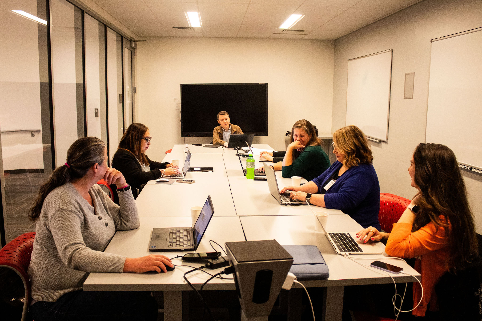 Seven participants sit around a conference table with laptops open, actively engaging in a meeting or workshop.