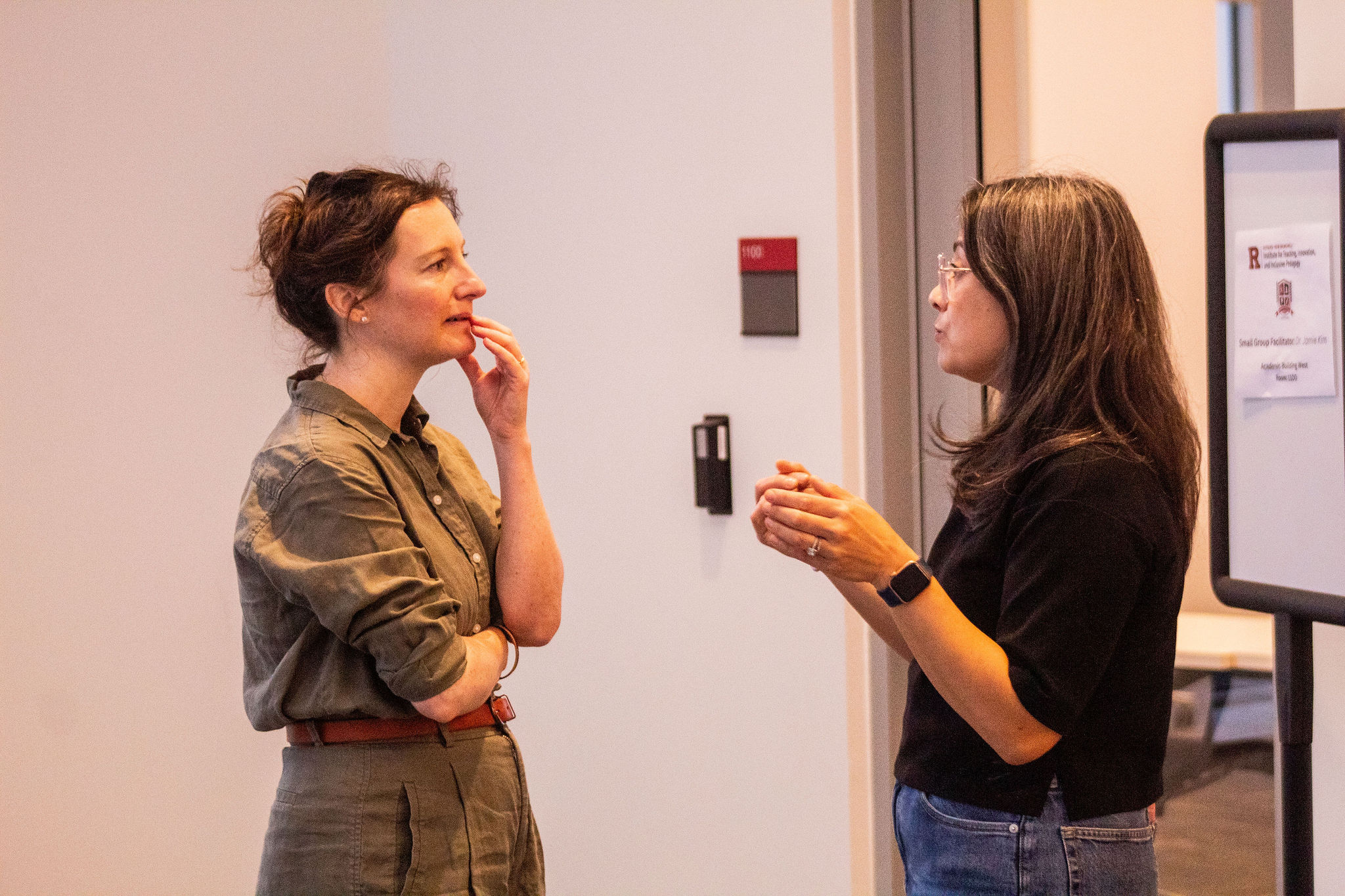 Two women stand in conversation near a wall and a sign, one speaking while the other listens thoughtfully with her hand on her chin.