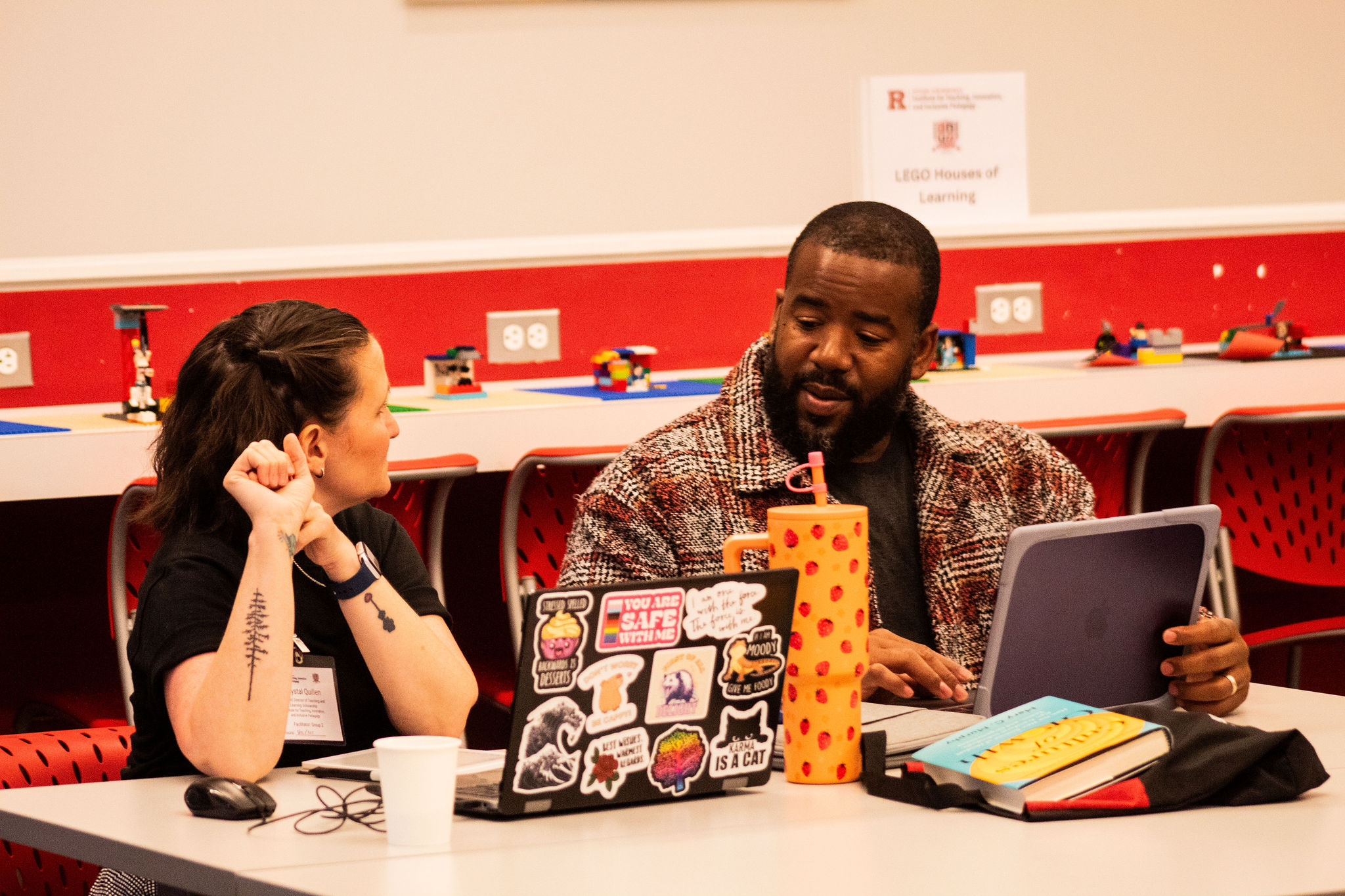Two colleagues sit at a table in a colorful room with LEGO models, chatting while working on laptops and reviewing materials.