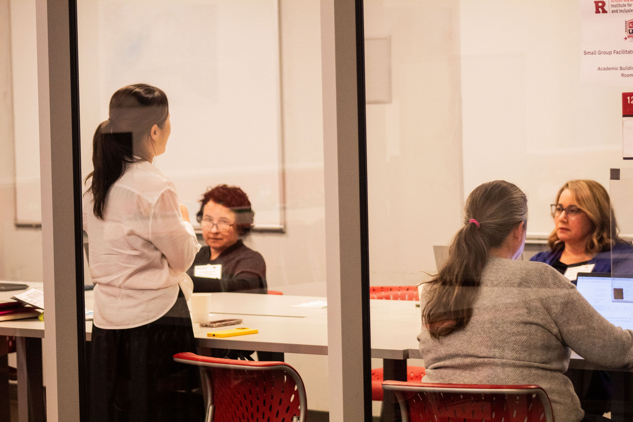 A woman leads a small group discussion in a classroom, as three seated participants listen and take notes.