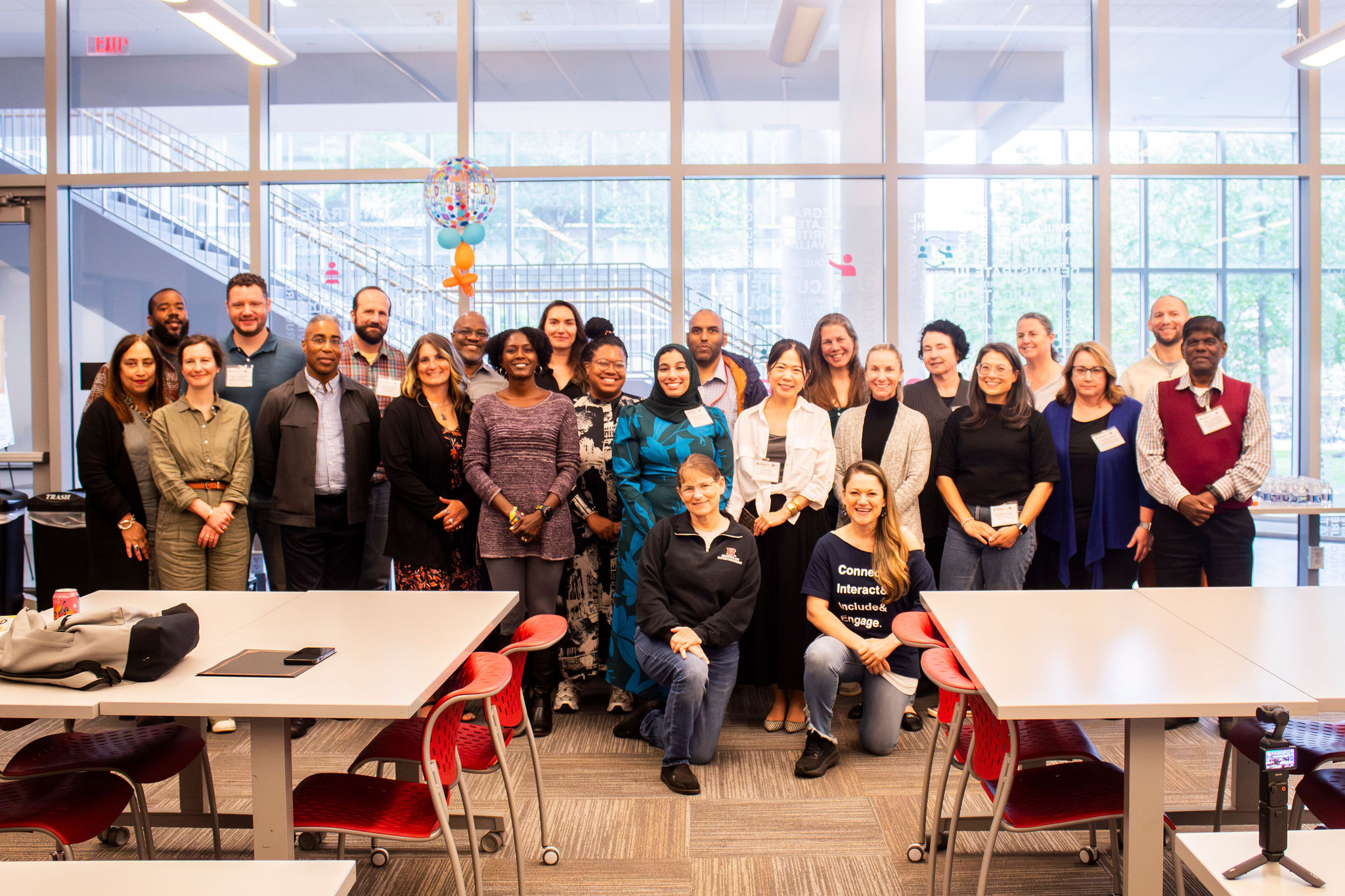 A group of faculty and staff posing together in a brightly lit room with large windows and red chairs, celebrating a professional event or workshop.
