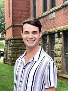 A man wearing a striped short-sleeve shirt smiles while standing outside in front of a brick building.