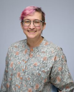 A person with short pink hair, glasses, and a patterned shirt smiles in front of a plain background.