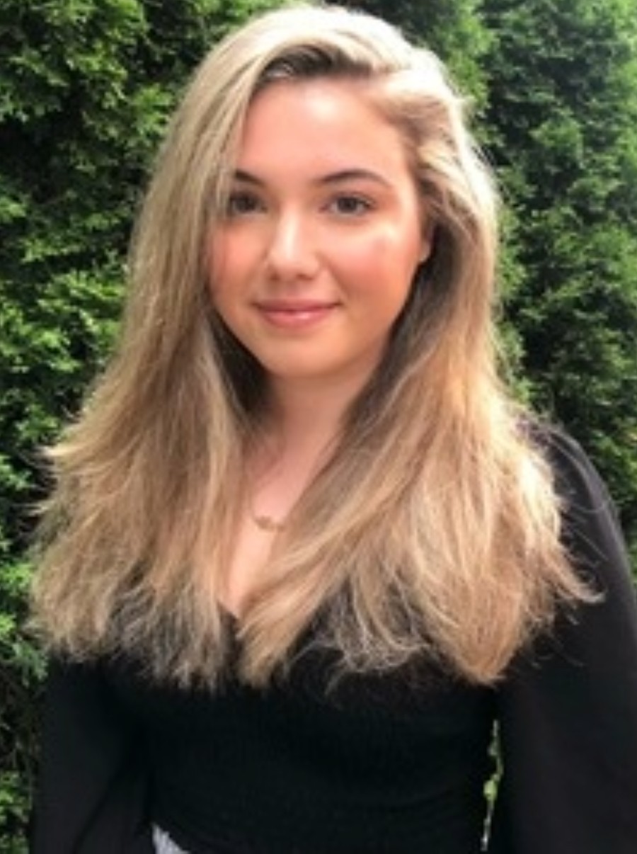 A young woman with long, light brown hair stands in front of tall green bushes, wearing a black top and smiling softly at the camera.