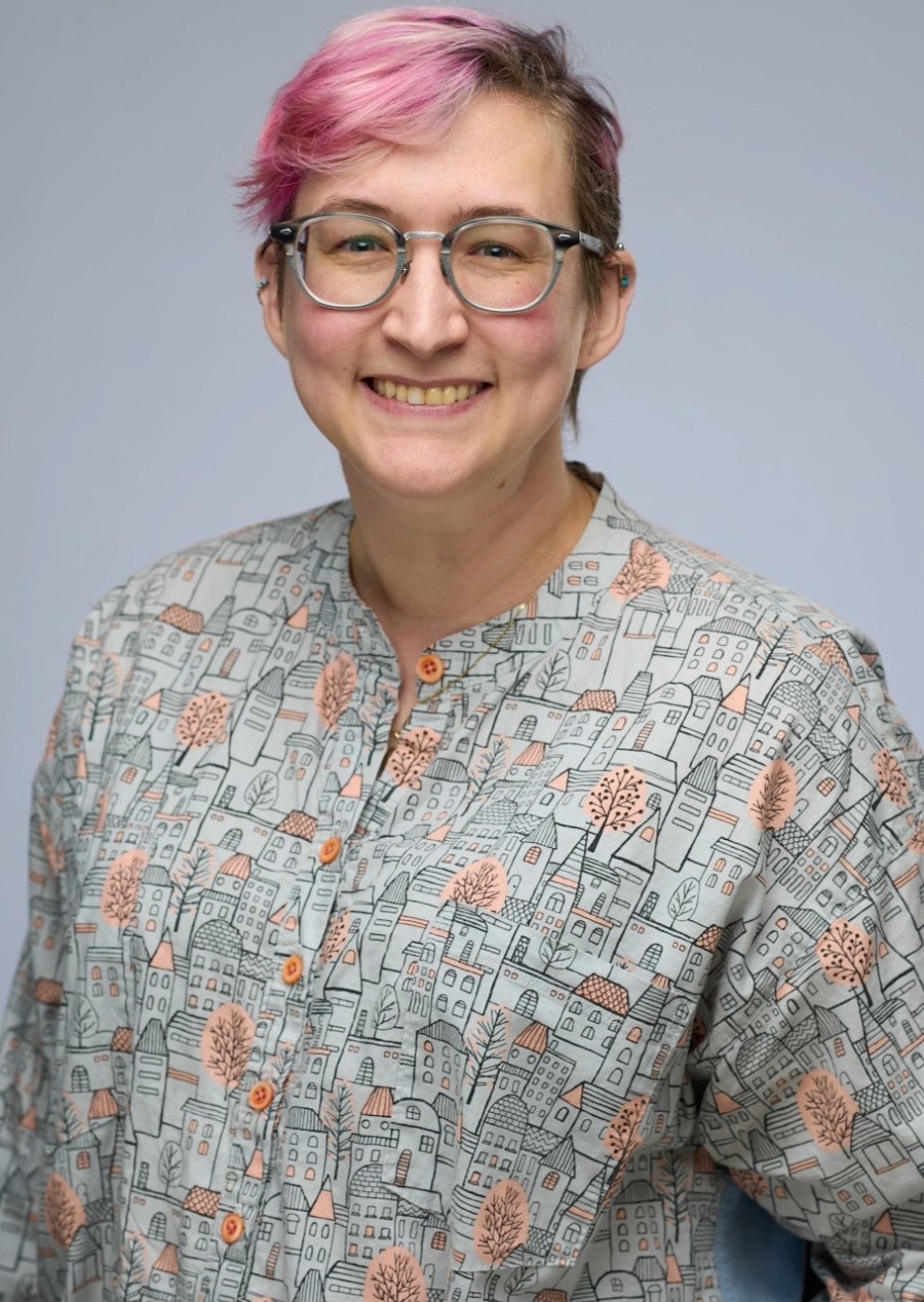 A person with short pink hair, glasses, and a patterned shirt smiles in front of a plain background.