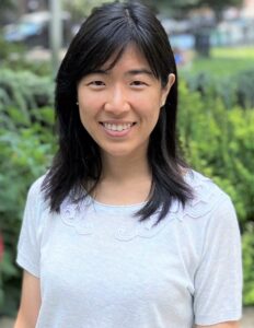 a woman with medium-length black hair wearing a white top, standing outdoors with a blurred garden background.