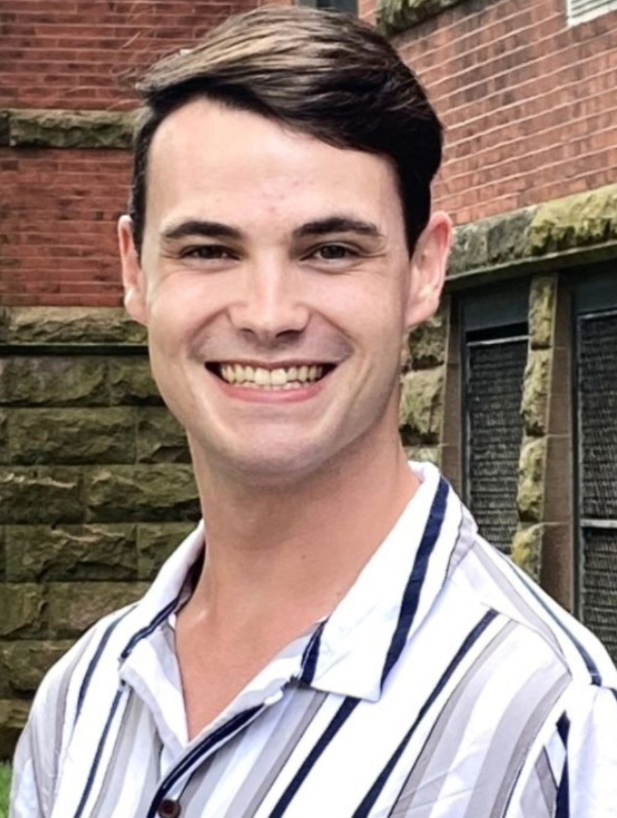 A man wearing a striped short-sleeve shirt smiles while standing outside in front of a brick building.