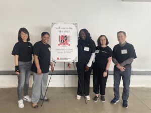 Five facilitators stand smiling beside a welcome sign for the May 2025 Course Design Institute at Rutgers University.