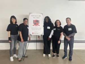 Five facilitators stand smiling beside a welcome sign for the May 2025 Course Design Institute at Rutgers University.