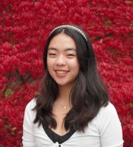 Headshot of undergraduate student fellow, Bethany Chen, wearing a white short and standing against background of red foliage.