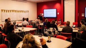A group of educators sit in groups at small tables, watching a presentation at the front of the room.