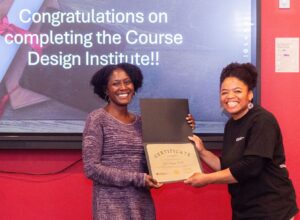 This photo shows two women smiling in front of a smartboard that reads "Congratulations on completing the Course Design Institute!!". One of the woman is handing a certificate to the other.
