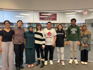 A group of eight students stand together in front of a glass wall, smiling. A bookshelf is shown behind them, and a sign above their heads reads "Rutgers University Press".