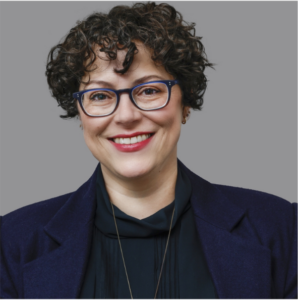 A headshot of a woman with short, curly, brown hair, wearing glasses and a blazer, smiling at the camera.