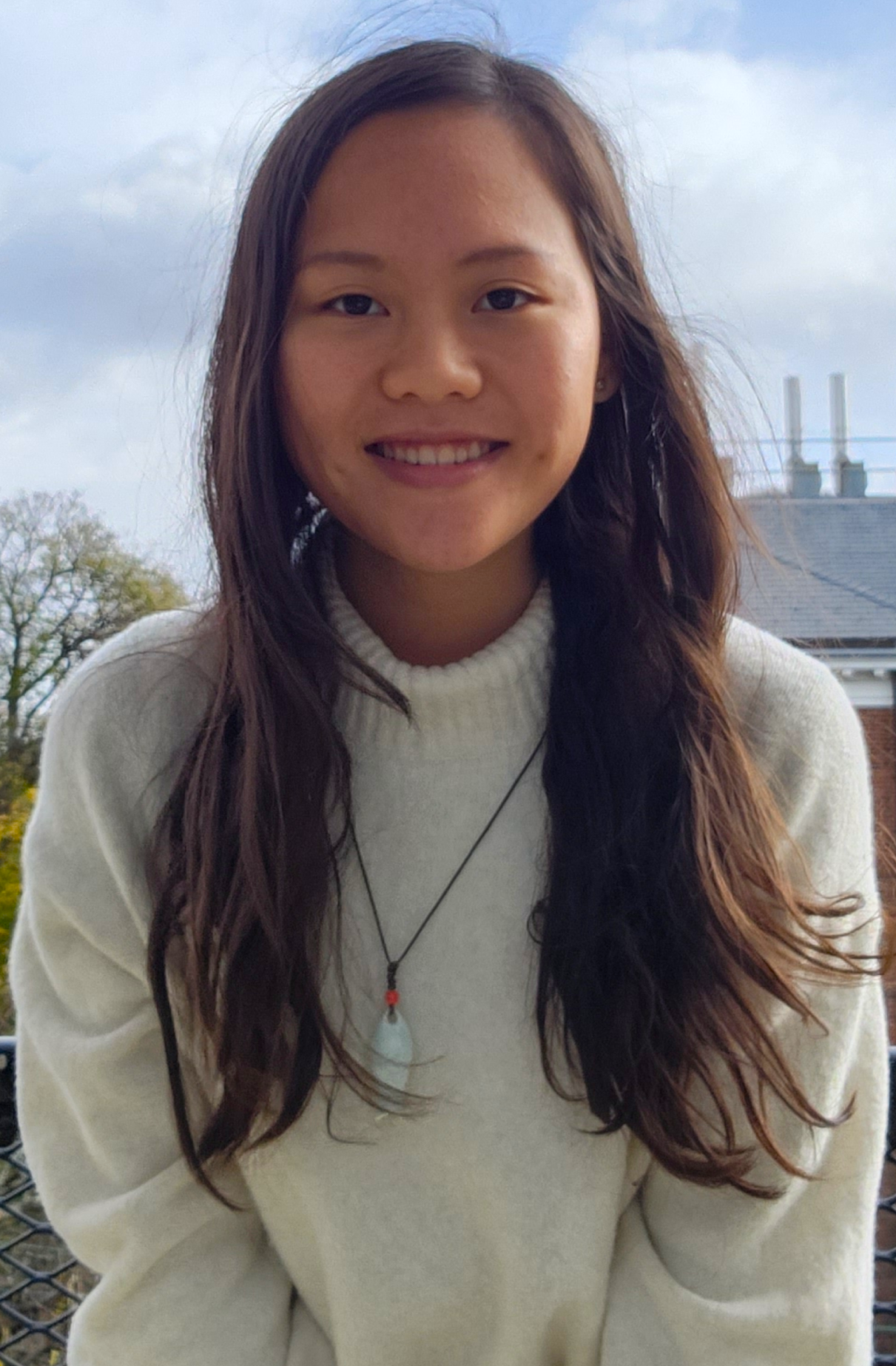 A young woman with brown hair in a white turtleneck shirt. In the background is a cloudy blue ski, some trees, and a brick building.
