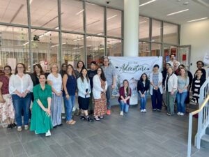 Twenty five individuals stand smiling for a group photo in a large, modern lobby, with large, floor-to-ceiling windows behind them. Behind them is a backdrop sign that reads "Let the Adventure Begin".