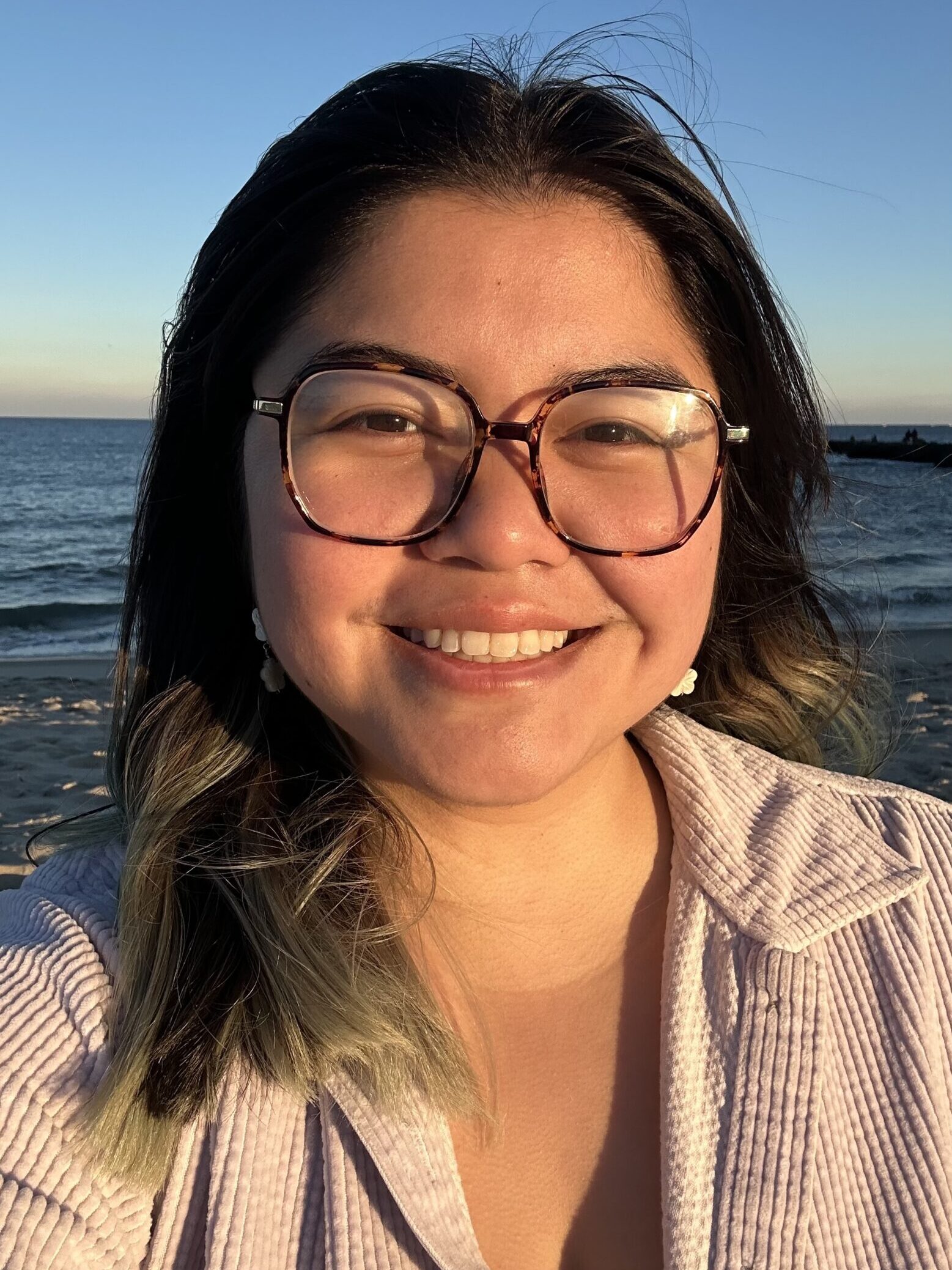 A headshot of a young woman wearing glasses and smiling. Behind her is a blue sky and the ocean.
