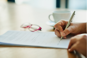 Image shows a desktop with a piece of paper, red glasses, and a mug. A person's hand is shown holding a pen, writing on the paper.