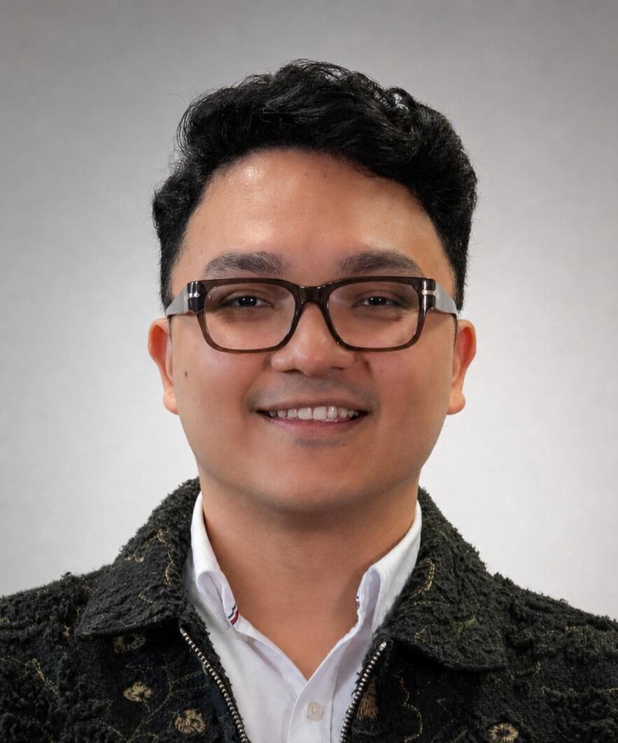 Headshot of a man in a white collared shirt and dark colored jacket, wearing glasses and smiling. The background of the photo is a gradient of gray-white.